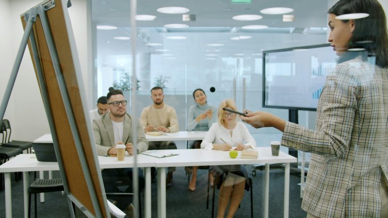 Presenter using a display screen and whiteboard while a seated team looks on in a meeting room.