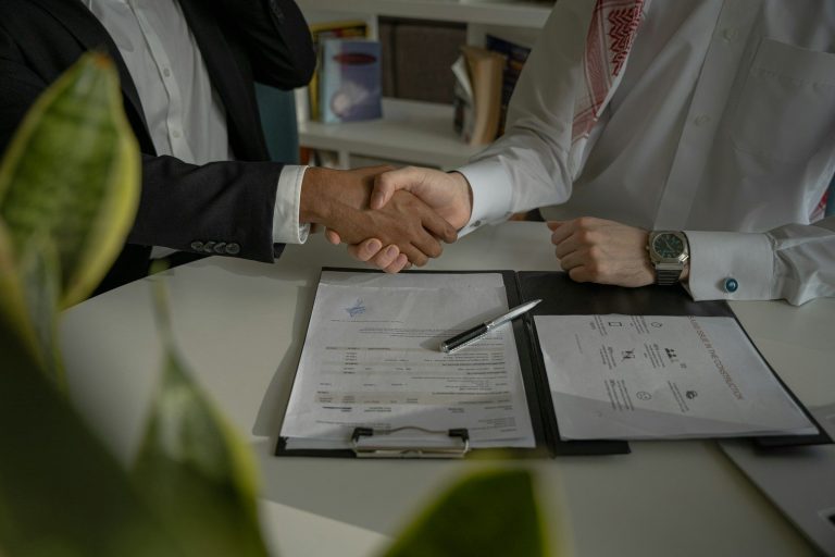 Two people shaking hands over a signed contract on a desk.