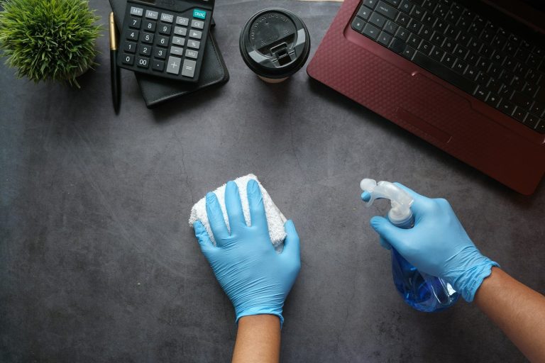 A gloved hand wipes a surface with a cloth while holding a cleaning spray.