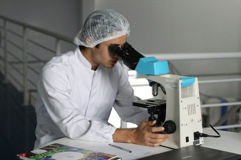 A scientist in a hairnet and white coat looks through a microscope in a laboratory.