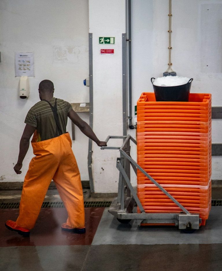 A worker in orange waterproof trousers pulls a trolley carrying a tall stack of orange crates inside a food handling facility.