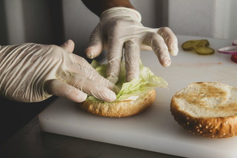 A person wearing clear disposable gloves assembles a burger with lettuce on a white cutting board.