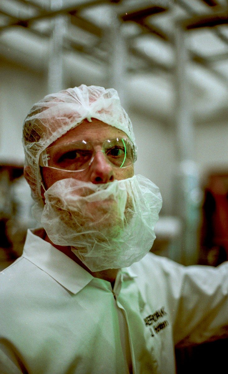 A food worker wearing a hairnet, beard cover, safety glasses and a white coat.