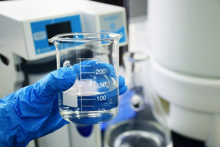 A gloved hand holds a glass measuring beaker containing a clear liquid.