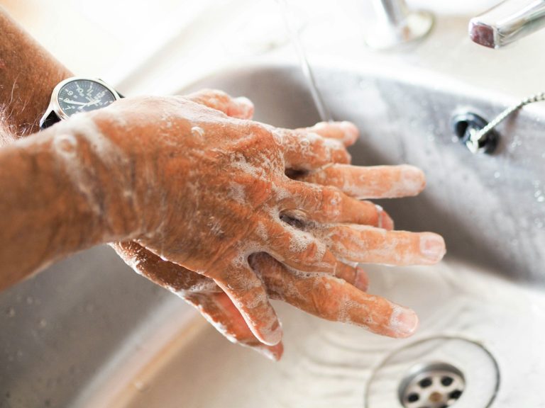 Hands covered in soap lather under running water at a sink during hand washing.