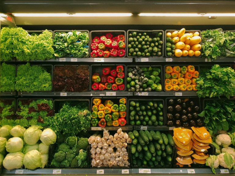 Fresh vegetables and salad greens are displayed in neat rows on lit shelves in a food retail store.