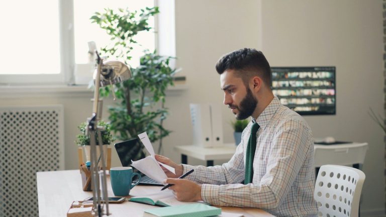 A man in a collared shirt reading a printed document at a desk beside a laptop.