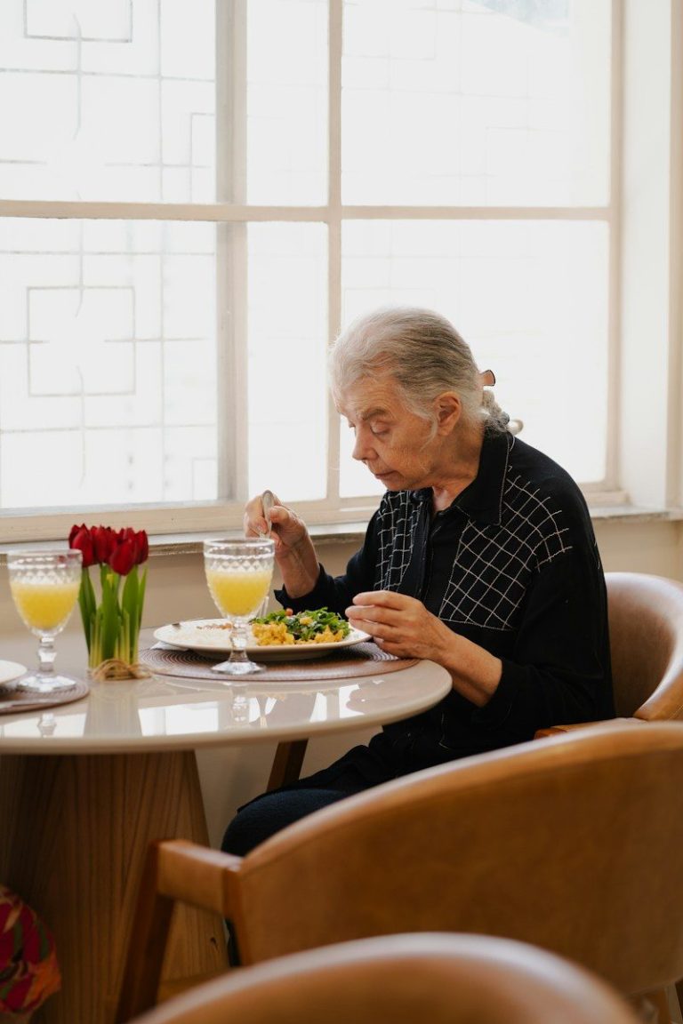 Older person eating a meal alone at a table beside a window