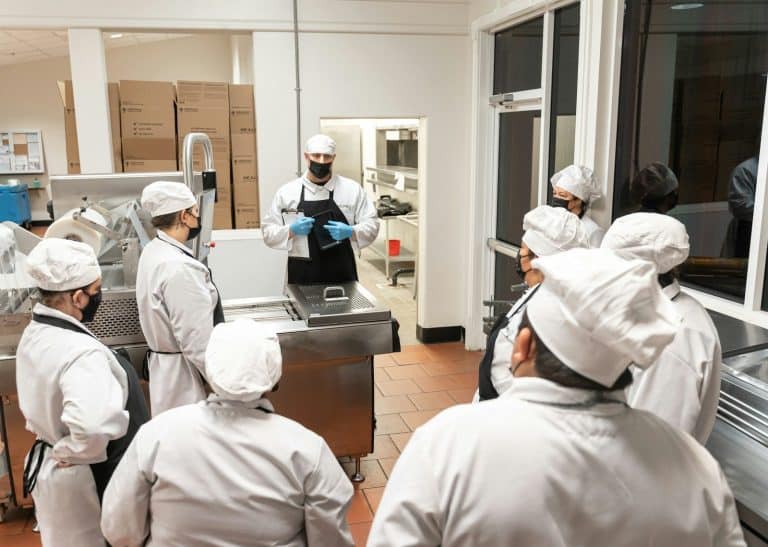 A supervisor in gloves briefs a group of staff in white coats and caps inside a commercial kitchen.