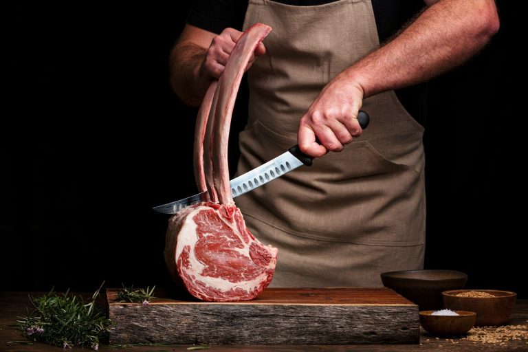 A butcher in a brown apron using a long knife to trim a rack of beef ribs on a wooden board.