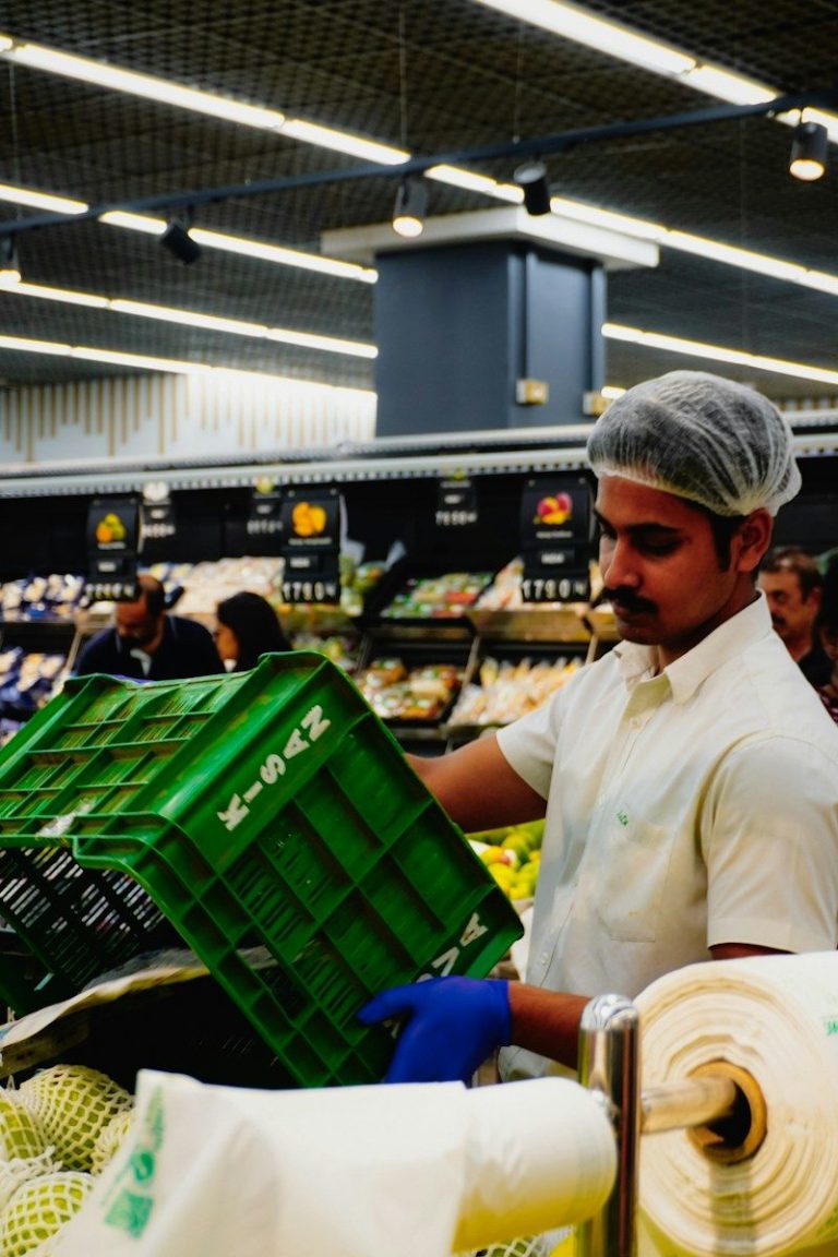 A food worker wearing a hairnet and white shirt lifts a green crate while working in a produce area.