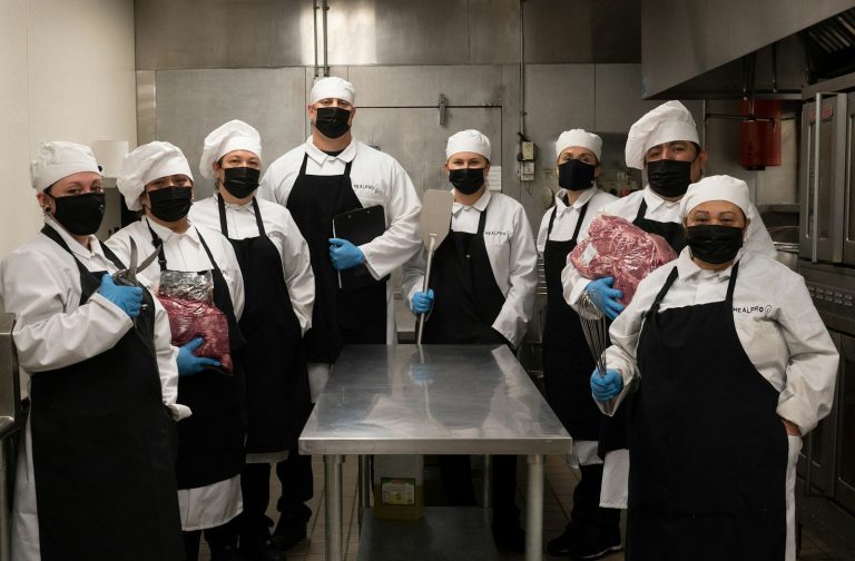 A group of kitchen staff in aprons, masks and gloves stand around a stainless steel bench holding food.