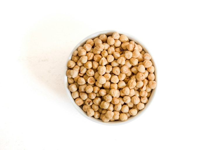 A white bowl of dry chickpeas on a plain white surface, representing pulse ingredients bought from approved suppliers.