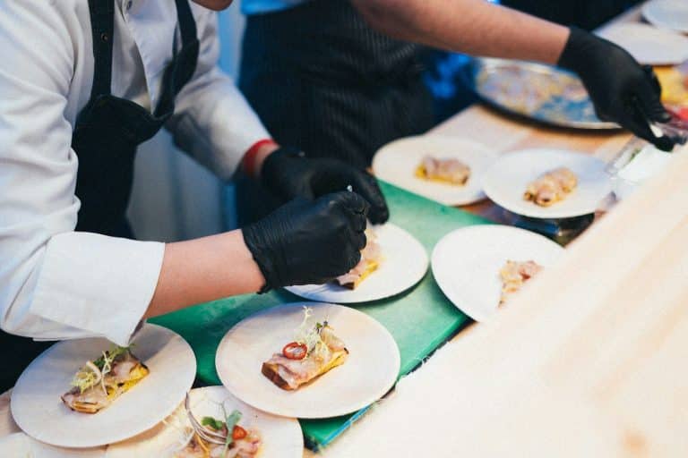 Food handlers wearing black gloves portion identical samples onto plates along a clean food service bench.