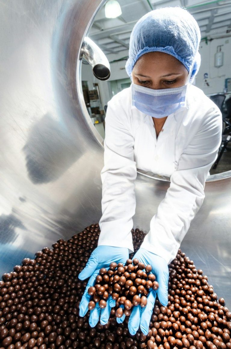 Worker in a hairnet, mask, and gloves handling chocolate coated products in a food factory