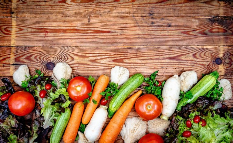 Fresh tomatoes, carrots, cucumbers, mushrooms and salad leaves arranged on a rustic timber surface.
