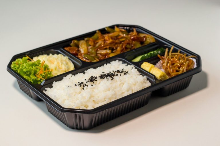 Black meal tray containing rice, stir fried vegetables and side dishes